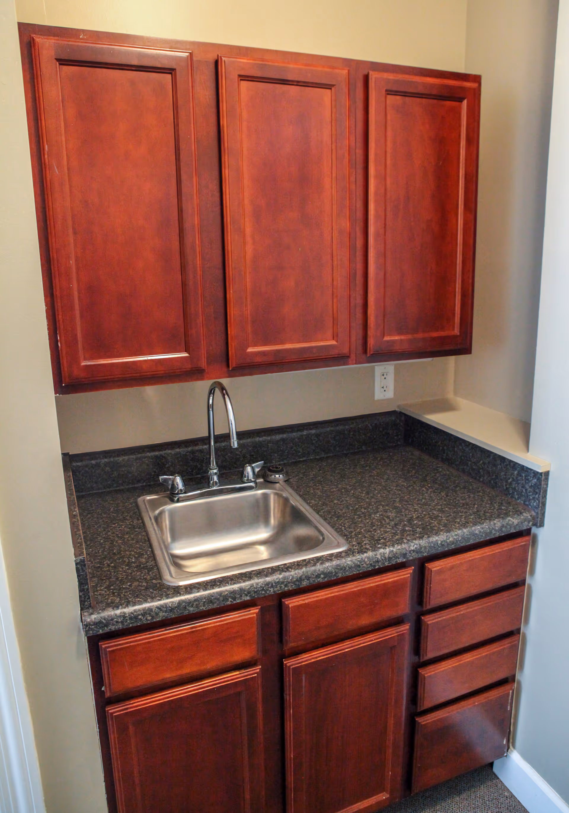 Small kitchenette with a stainless steel sink, dark speckled countertop, and cherry wood upper and lower cabinets.