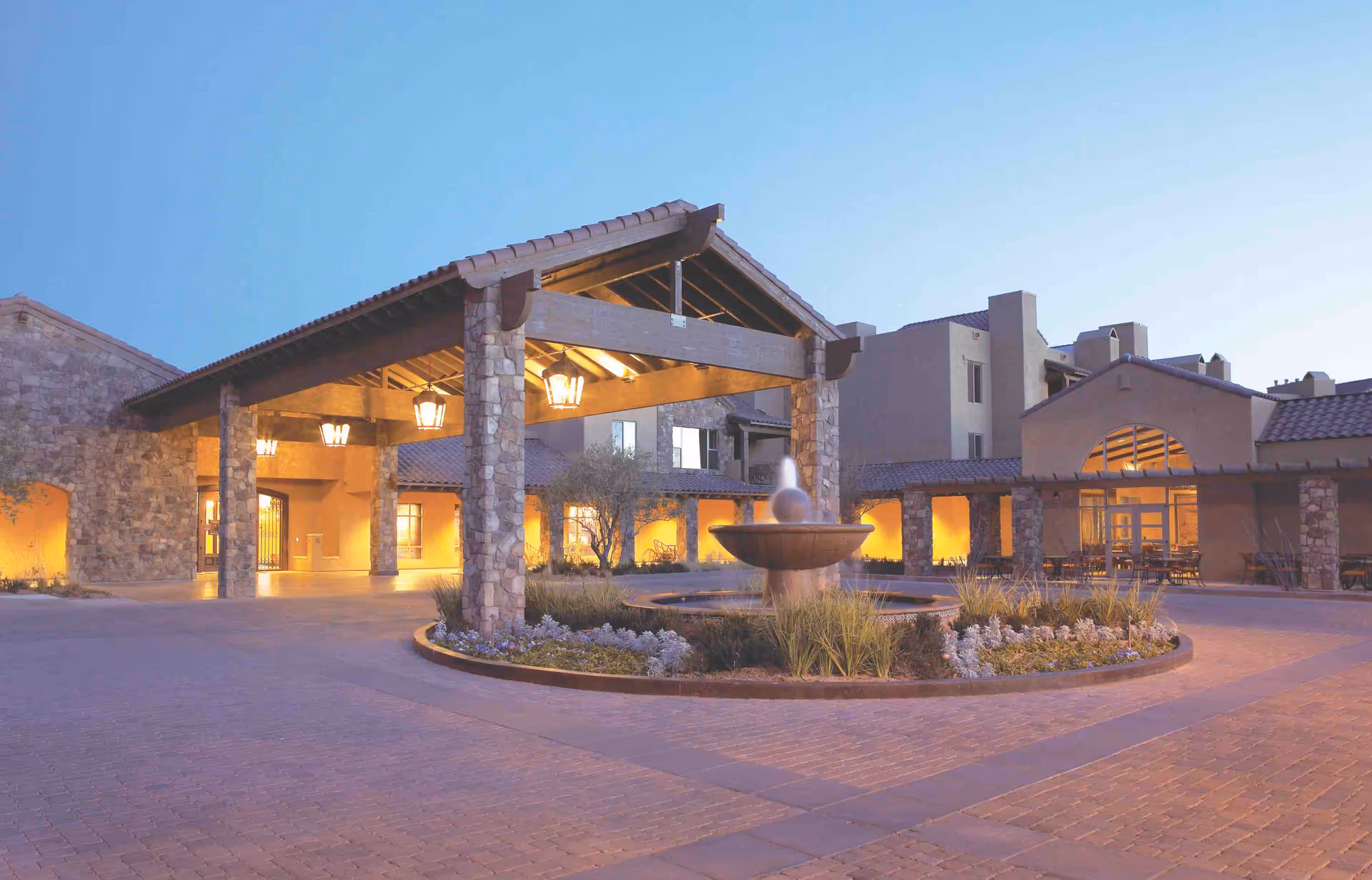Stone entry porte-cochère, fountain, and warm lights at Splendido senior living exterior.