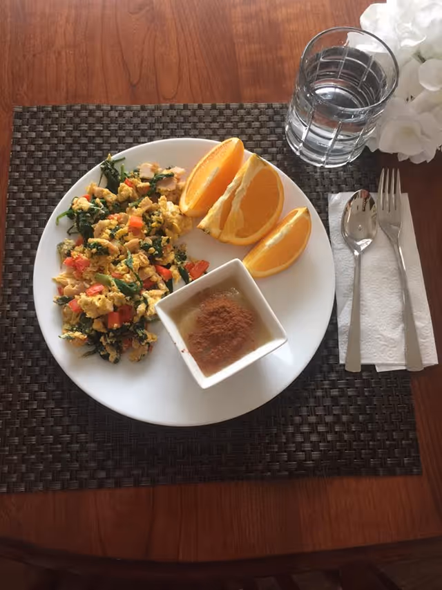 A white plate on a woven placemat with scrambled eggs mixed with vegetables, three orange wedges, and a small square dish topped with cinnamon. Next to the plate is a glass of water, a spoon, and a fork on a folded napkin, with white flowers in the background.
