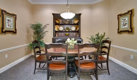 A small dining room with a wooden table and six chairs, a china cabinet displaying plates, potted plants, framed artwork, and a central chandelier.