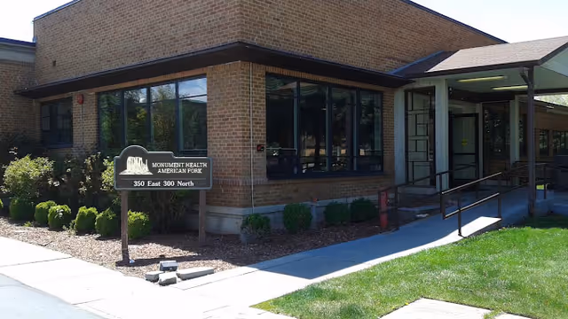 Exterior view of a brick building with large windows and a ramp leading to the entrance. A sign in front reads 'Monument Health American Fork, 350 East 300 North'. There are small bushes and landscaping around the building.