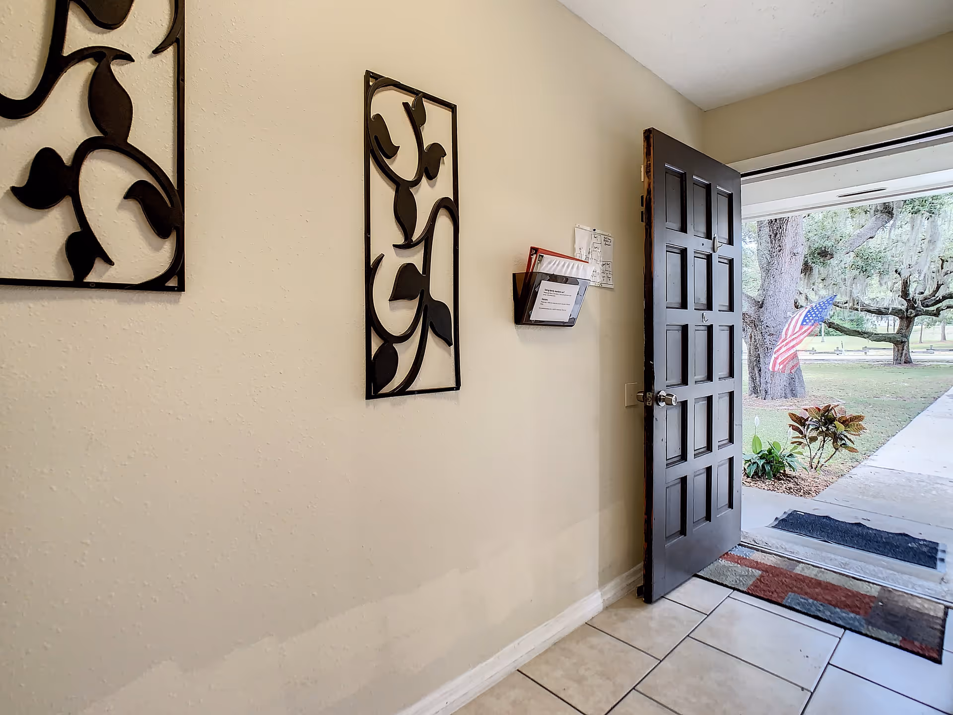 View of an open front door leading outside to a walkway and a lawn with trees and an American flag. Inside, there is a tiled floor, a wall with two decorative black metal wall hangings, and a wall-mounted holder with papers.
