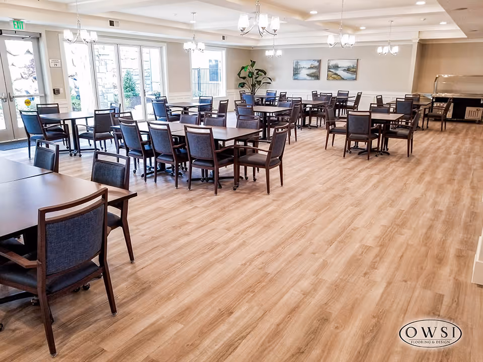 A spacious dining room with multiple wooden tables and chairs arranged neatly on a light wood floor. The room has large windows and glass doors allowing natural light to enter. There are chandeliers hanging from the ceiling, two landscape paintings on the wall, and a large plant in the corner. The room appears clean and well-lit.