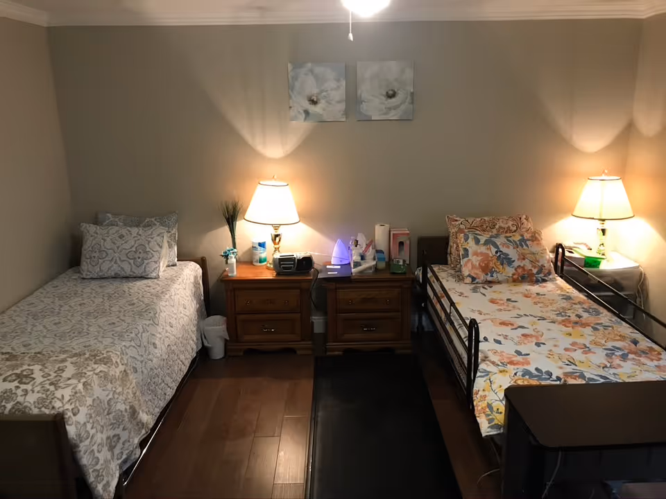 A cozy bedroom with two single beds separated by two wooden nightstands. Each nightstand has a lamp, and various personal items are placed on top. The bed on the left has a patterned gray and white bedspread with matching pillows, while the bed on the right has a floral bedspread with multiple pillows. Two floral paintings hang on the beige wall above the nightstands. The floor is wooden with a dark rug between the beds.