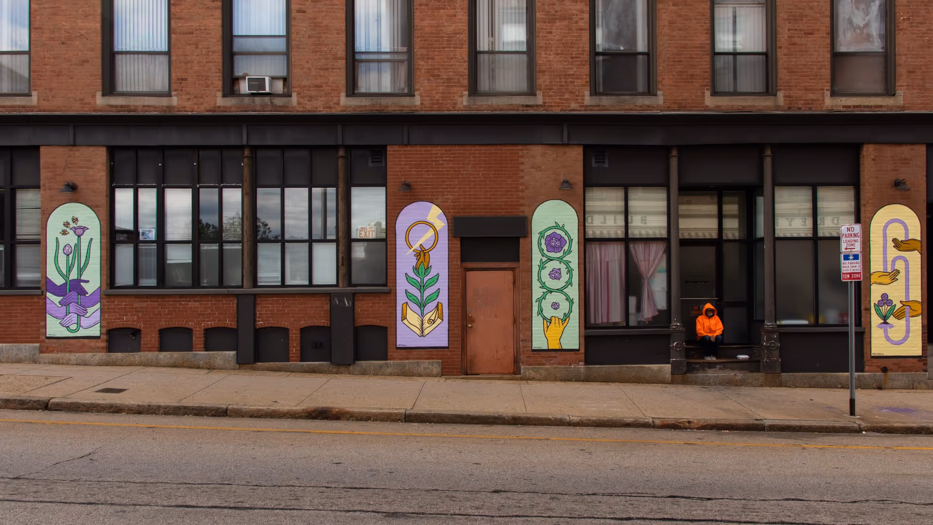 Exterior view of a brick building with large windows and colorful murals painted on panels between the windows. A person wearing an orange hoodie is sitting on the steps near the entrance. There is a 'No Parking Loading Zone' sign on the sidewalk.