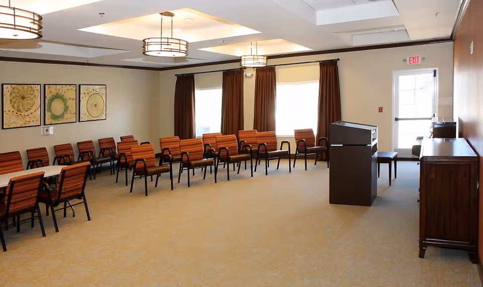 A spacious room with multiple rows of orange-striped chairs arranged in a semi-circle and around tables, beige carpet, three large windows with brown curtains, three ceiling light fixtures, and framed artwork on the wall. There is a dark wooden podium and a cabinet near the exit door.