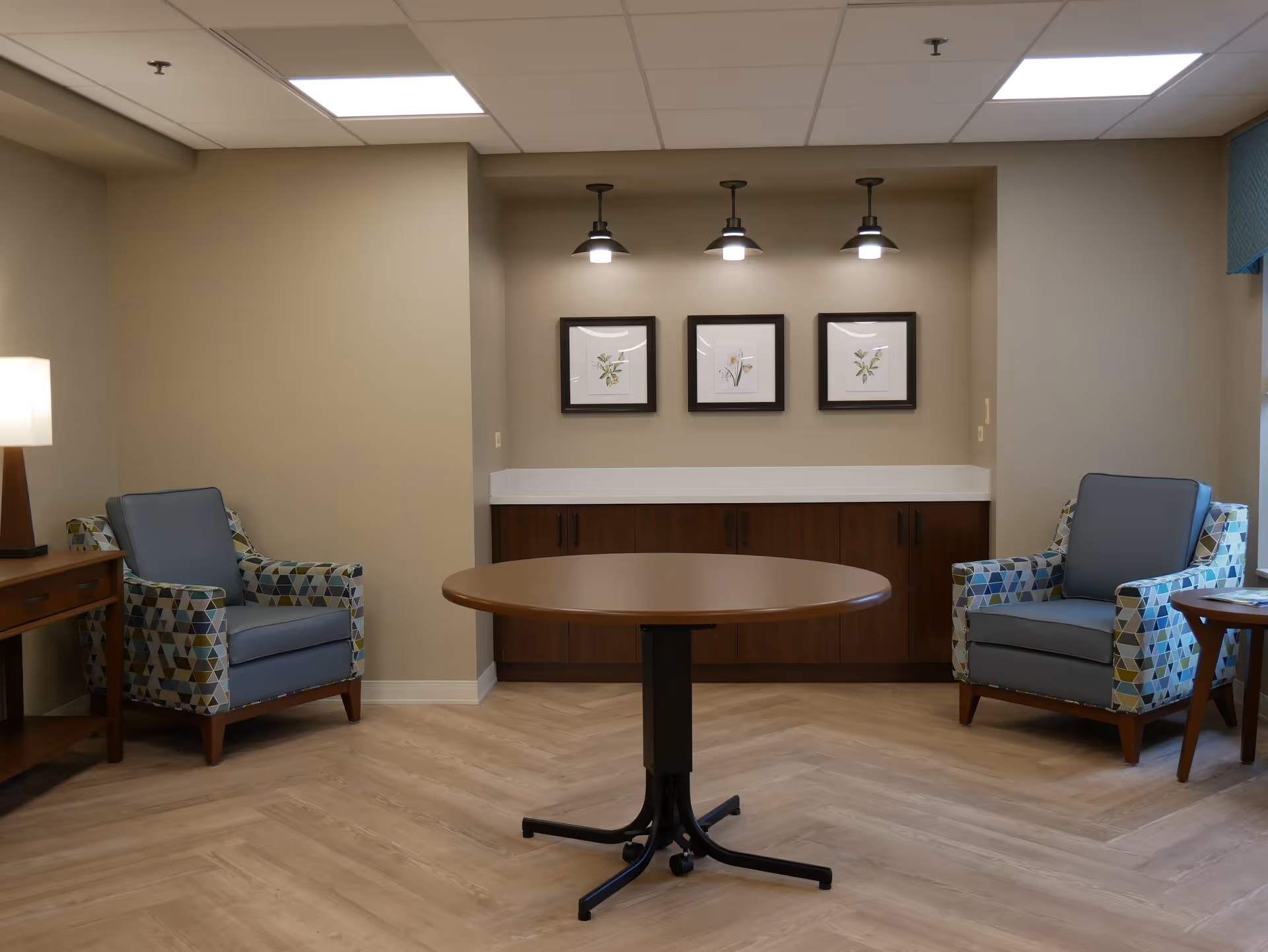 A cozy sitting area in a nursing and rehabilitation facility featuring two patterned armchairs with blue cushions placed on either side of a round wooden table. The background shows a wooden cabinet with a white countertop, above which are three framed botanical prints illuminated by three small pendant lights. The room has light-colored walls, wood-patterned flooring, and a ceiling with recessed lighting.