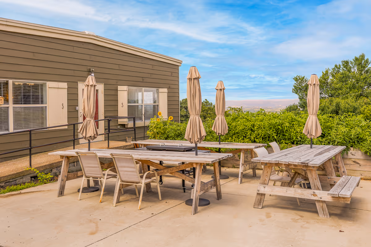 Outdoor patio area with wooden picnic tables, chairs and closed umbrellas beside a low building and green shrubbery under a blue sky.