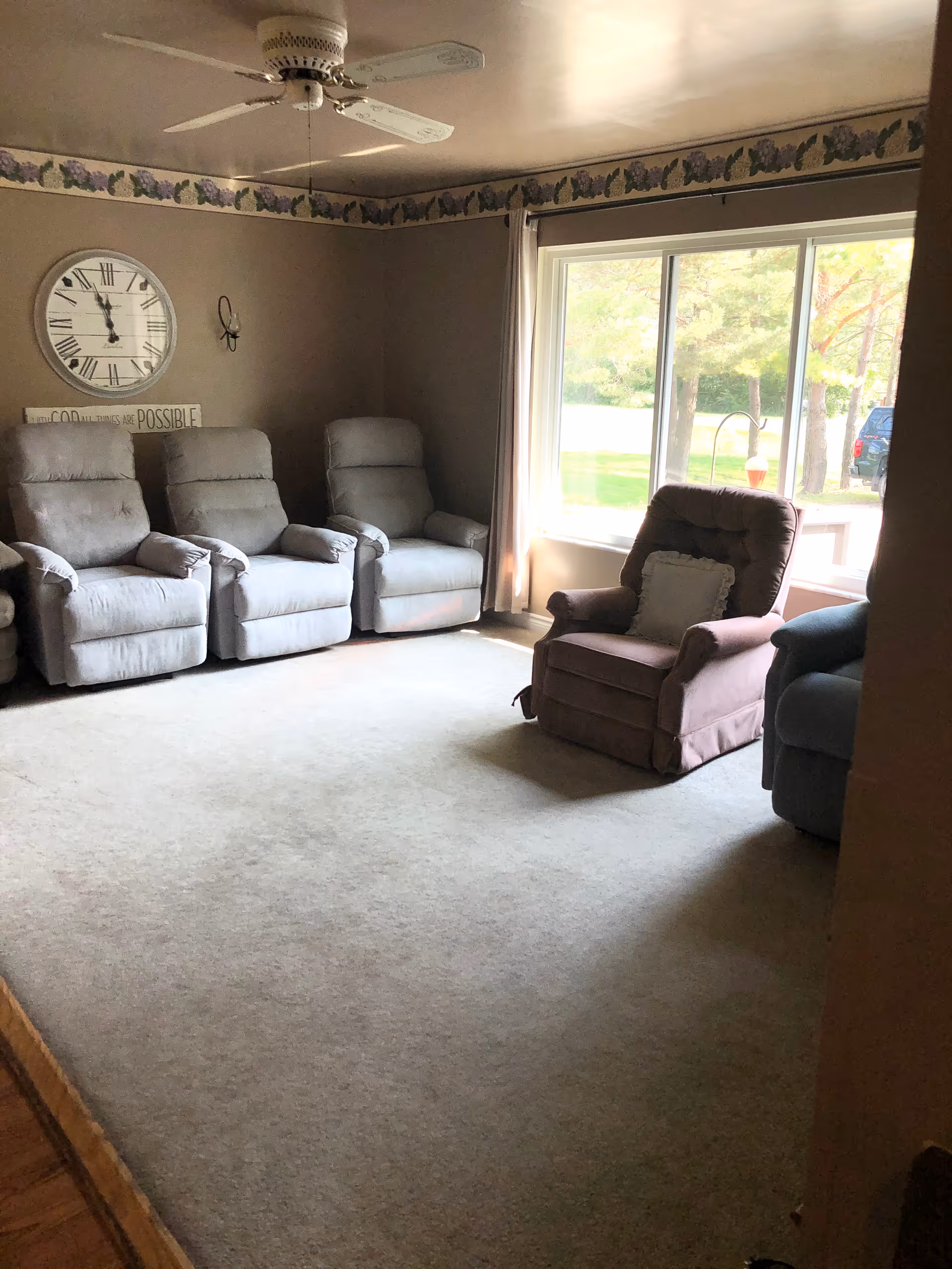 A living room with four gray recliner chairs and one brown recliner chair arranged around the room. There is a large window letting in natural light, a ceiling fan, a wall clock, and a decorative border near the ceiling.