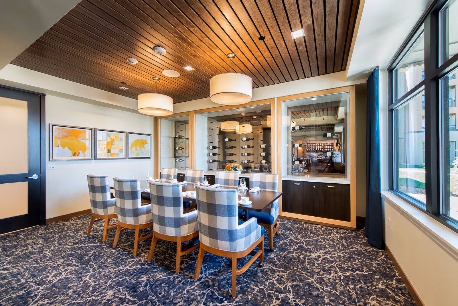A dining room with a rectangular table set for eight people with plaid upholstered chairs. The room features a wooden slatted ceiling with two large pendant lights, a patterned carpet, and large windows letting in natural light. On one wall, there are three framed animal artworks, and a glass-enclosed wine display is visible.
