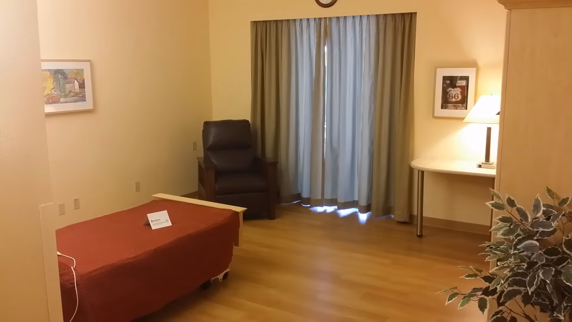 A simple bedroom with a single bed covered in a red blanket, a dark brown recliner chair, a small table with a lamp, framed pictures on the walls, and a window with closed curtains. There is a plant partially visible in the foreground.