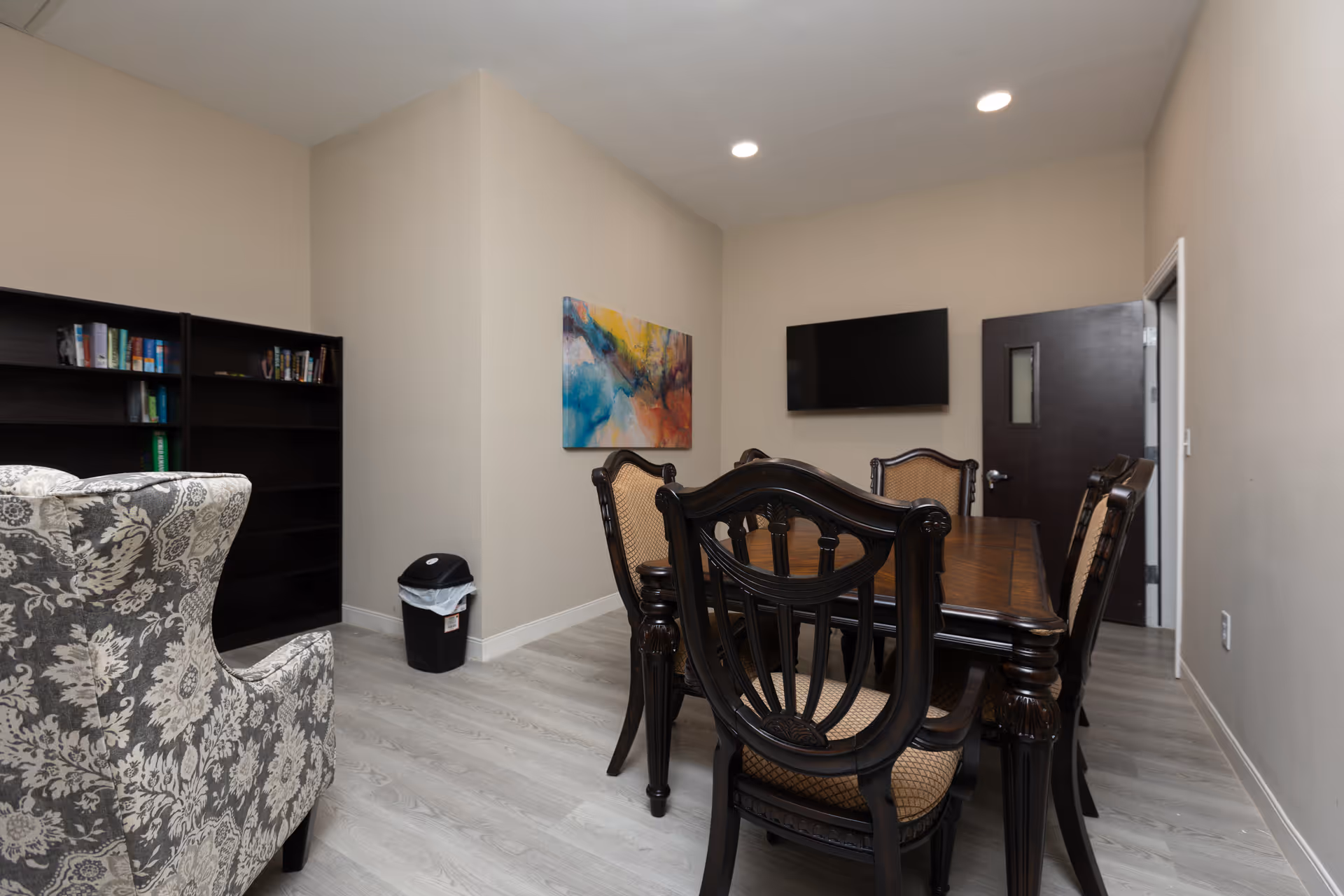 A dining/meeting room with a dark wooden table and chairs, an upholstered armchair, bookshelf, wall-mounted TV and abstract painting.