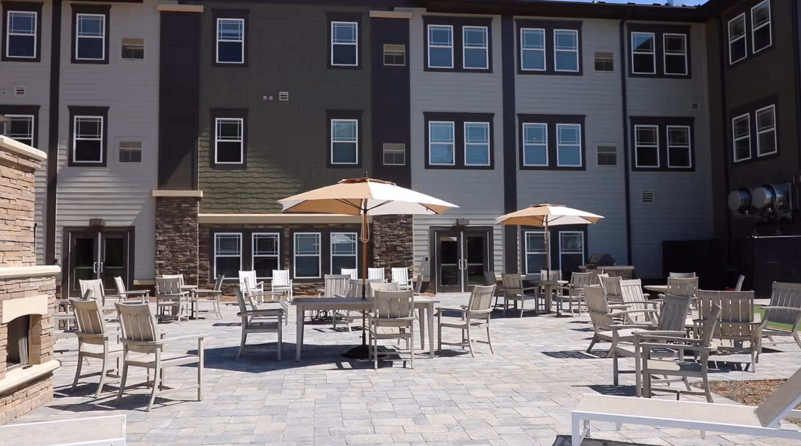 Outdoor patio area with multiple wooden chairs and tables, some shaded by large beige umbrellas, in front of a multi-story building with many windows. There is a stone fireplace on the left side of the patio.