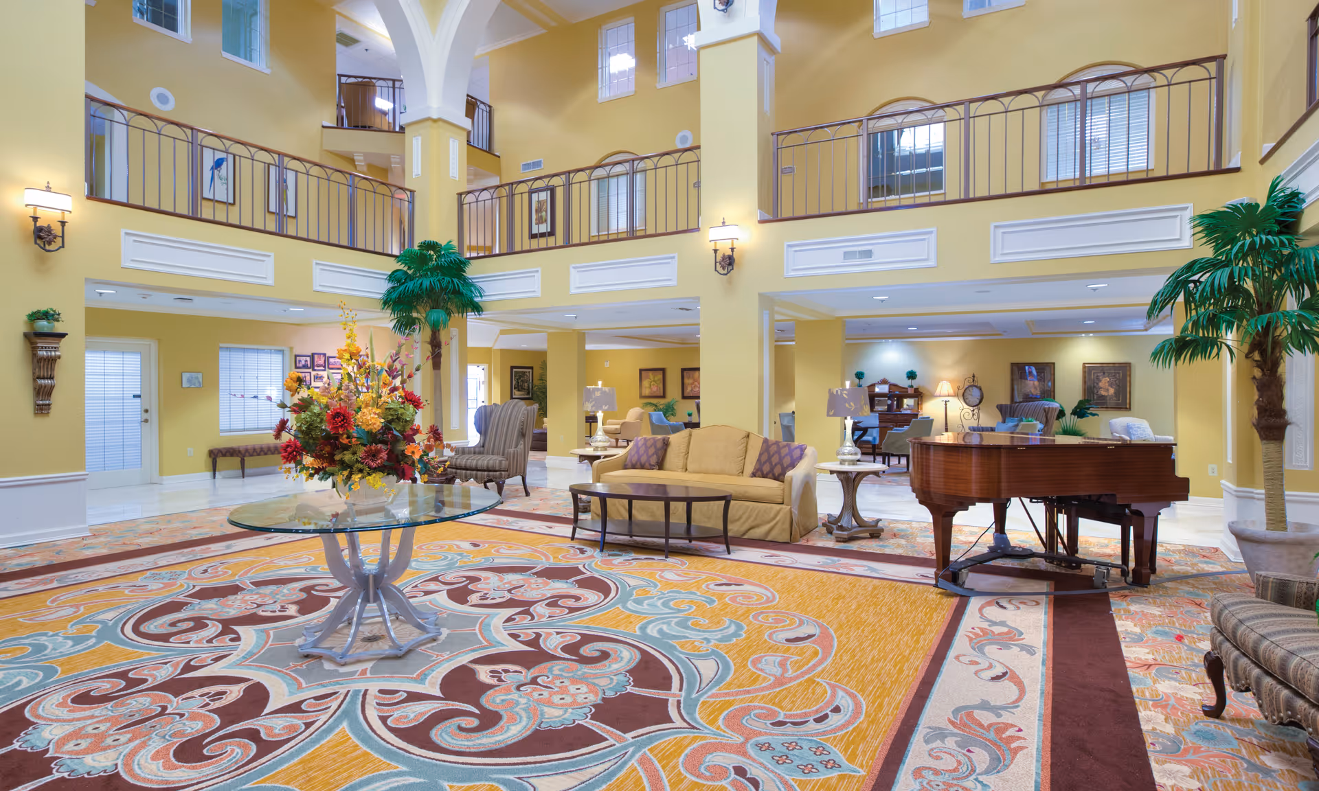Spacious two-story senior living lobby with a grand piano, seating area, central glass table with a floral arrangement, and a large patterned rug.