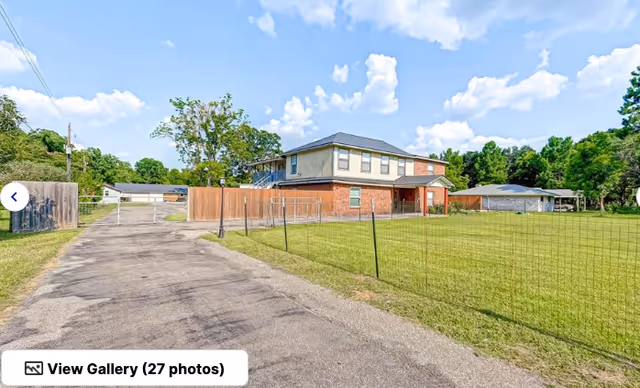 Two-story brick and siding assisted living building with a fenced grassy yard and a long driveway under a partly cloudy sky.