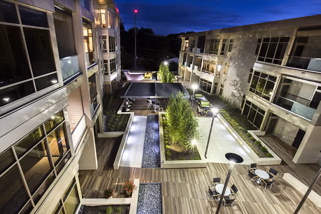 Nighttime view of a modern senior-living courtyard with illuminated walkways, seating areas, planters, and surrounding multi-story buildings.