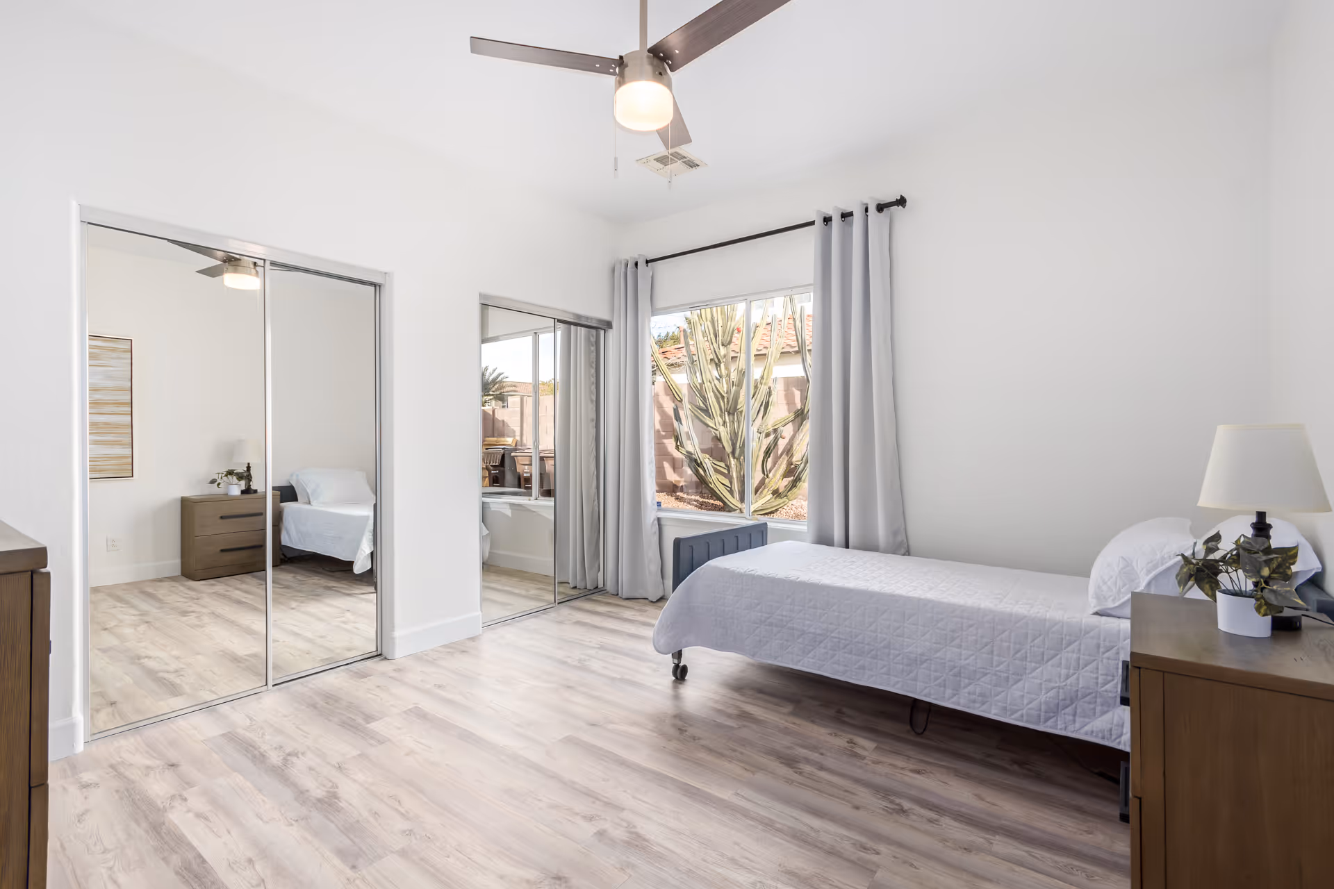 A bright and clean bedroom with light wood flooring, a single bed with white bedding, a wooden nightstand with a lamp and a plant, mirrored sliding closet doors, a large window with gray curtains showing an outdoor view with a cactus, and a ceiling fan with a light.