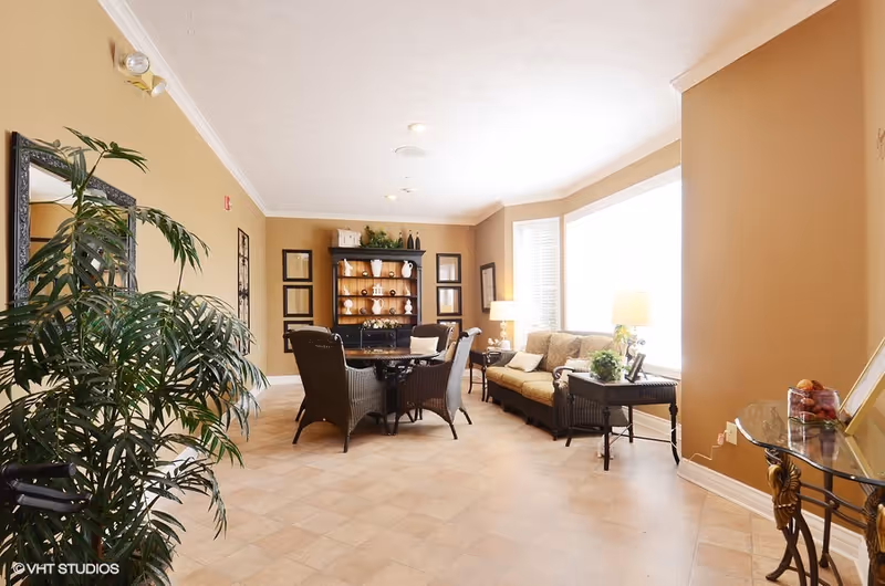 A bright and spacious living room area with beige walls and tiled floor. The room features a black wooden cabinet with decorative items, a round table surrounded by four wicker chairs, a beige sofa with cushions, two side tables with lamps, and a large green plant near a mirror on the left wall. Large windows on the right side allow natural light to fill the room.