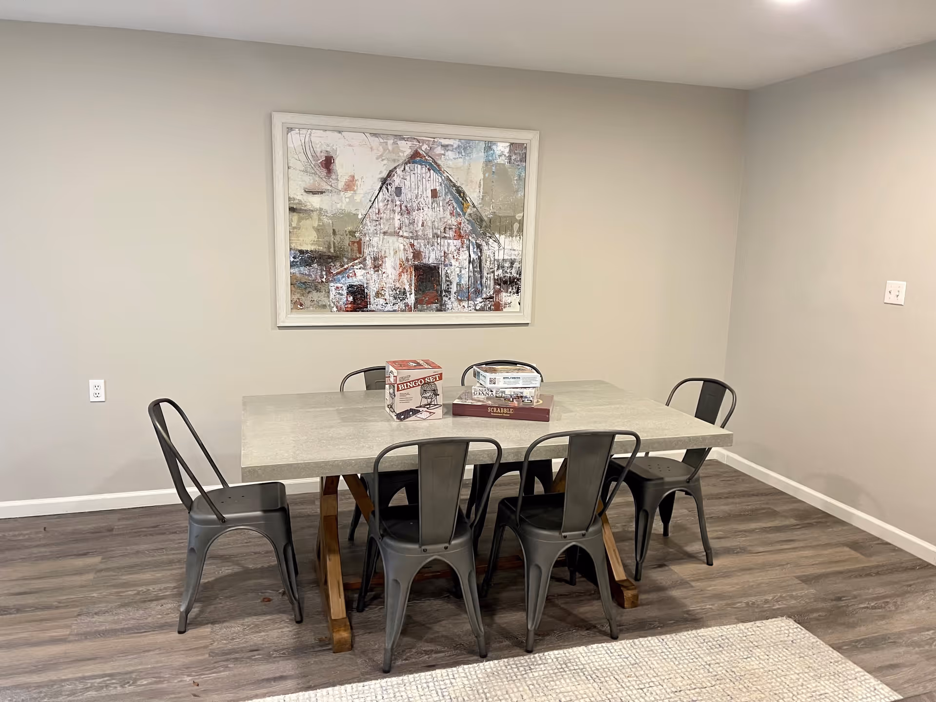A simple dining area with a rectangular table and six metal chairs. On the table are a bingo set, a Scrabble game, and a clear container. A large framed painting of a barn hangs on the wall behind the table. The room has light-colored walls and wood flooring.