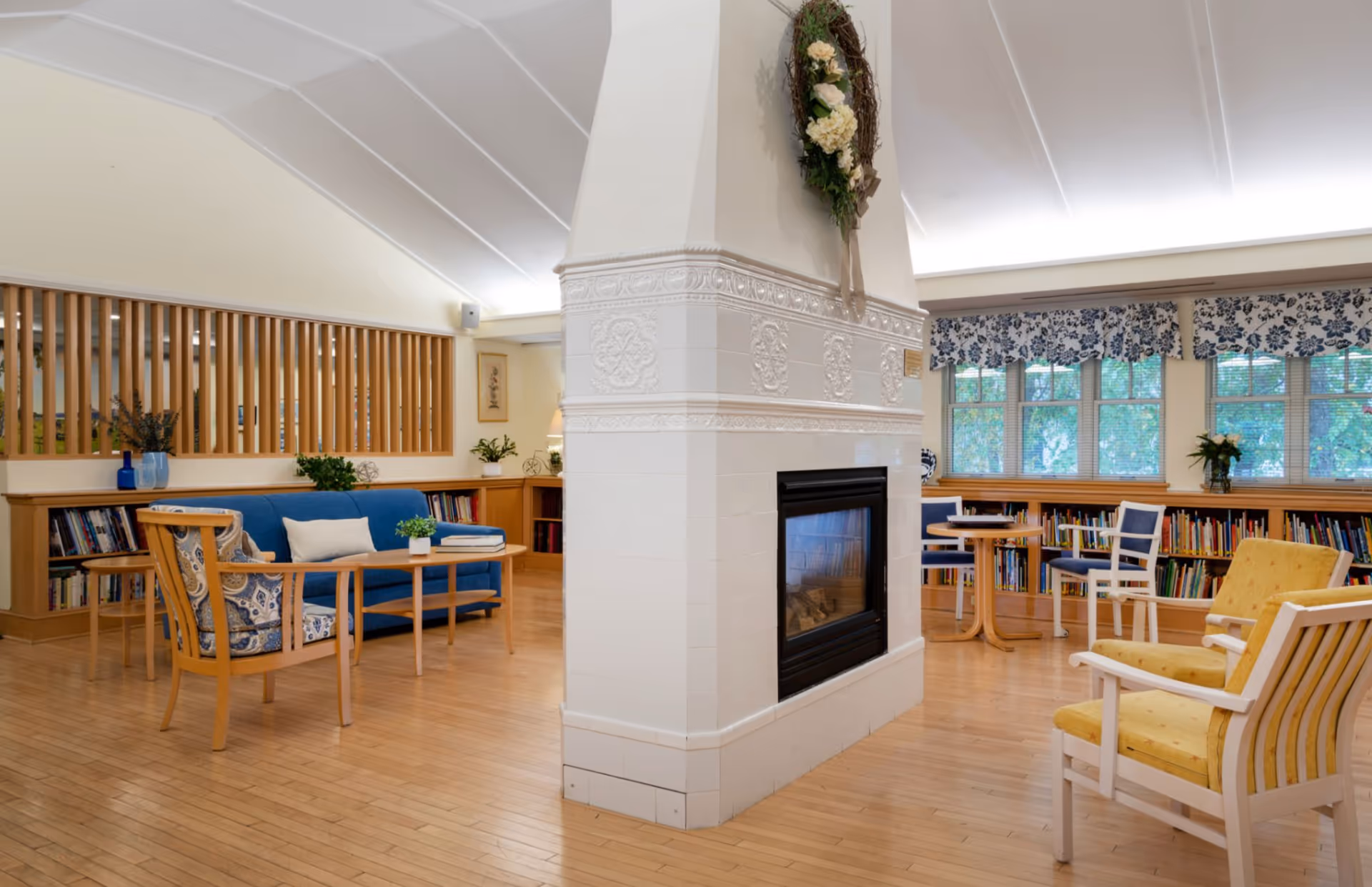 Cozy communal living room with a central white tiled fireplace, seating, bookshelves and large windows.
