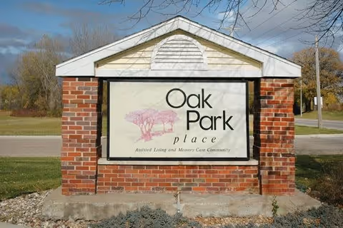 A brick and white wooden sign structure displaying the name 'Oak Park place' with a pink tree graphic and the text 'Assisted Living and Memory Care Community' underneath, situated outdoors with grass, trees, and a road in the background.