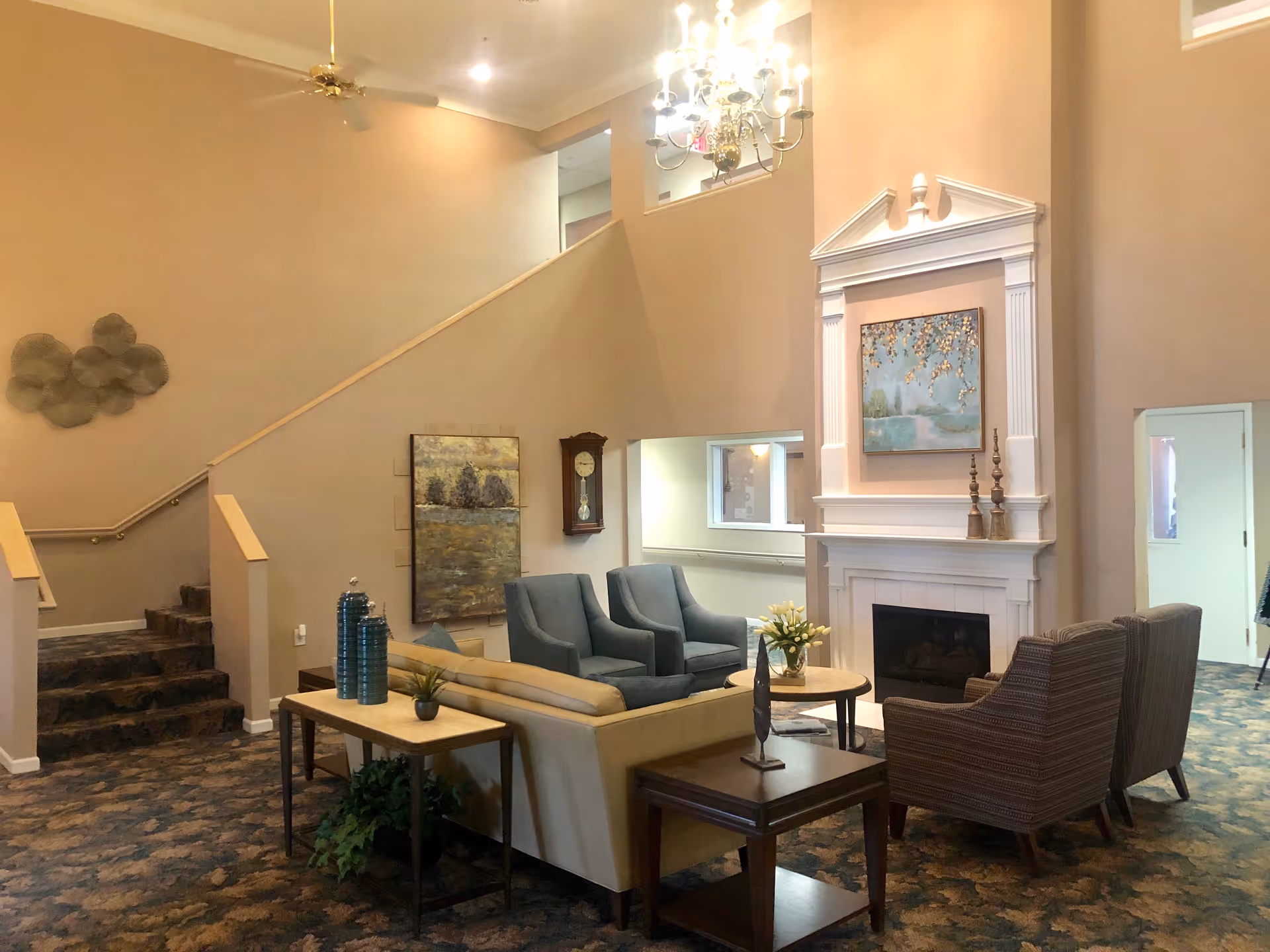 A cozy living room area in a retirement community featuring a beige sofa, two blue armchairs, and two striped armchairs arranged around a small round table with a vase of flowers. There is a white fireplace with a painting above it, a grandfather clock on the wall, and a staircase leading to an upper floor. The room has warm beige walls, a ceiling fan, a chandelier, and patterned carpet.