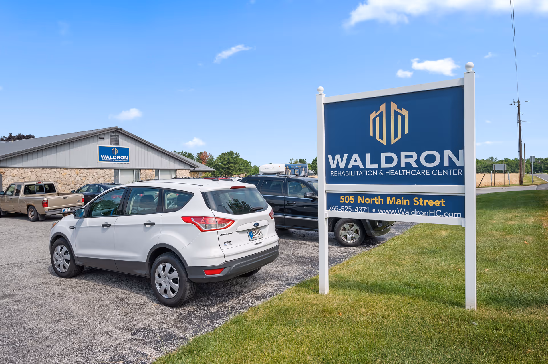 Parking lot outside Waldron Rehabilitation and Healthcare Center with several parked vehicles and a large blue sign displaying the facility's name, address, phone number, and website. The building is visible in the background under a partly cloudy sky.