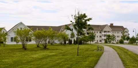 A large senior living facility building with white siding and multiple windows, surrounded by green grass, small trees, and a paved walkway with a bench alongside it under a partly cloudy sky.