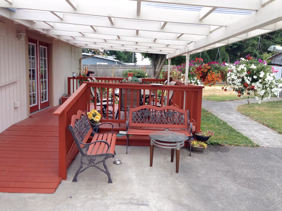 Outdoor patio area with red wooden benches and a glass-top table under a white pergola. Hanging flower baskets with colorful blooms are suspended from the pergola. The patio is adjacent to a building with red-framed glass doors and a red wooden ramp. A concrete pathway and grassy area are visible in the background.