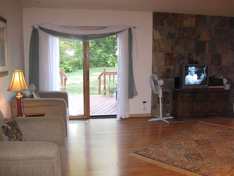 A cozy living room with two beige armchairs, a wooden side table with a lamp, a sliding glass door with white and green curtains leading to a wooden deck and green yard outside, a standing fan, a wooden cabinet with a small TV playing a black and white show, and a stone accent wall behind the TV. The floor is wooden with a patterned area rug partially visible.