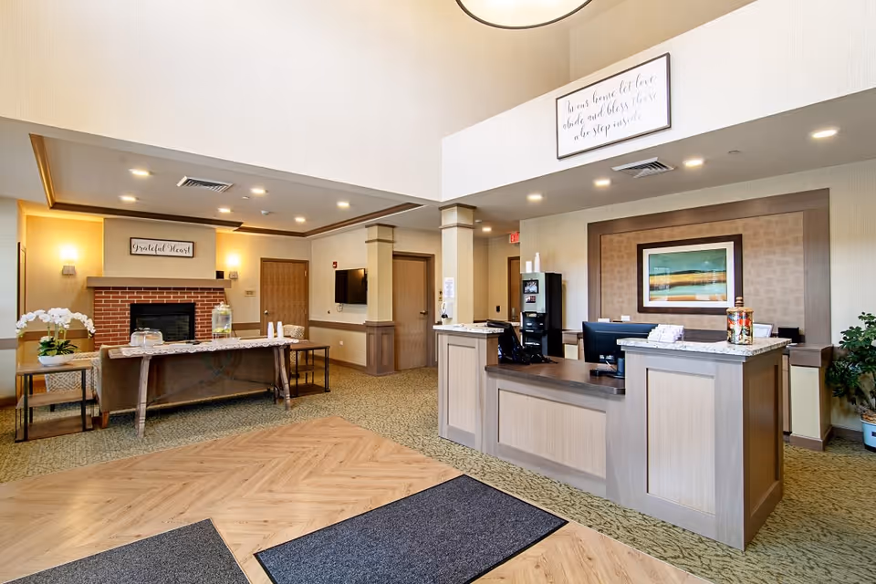 A spacious and well-lit reception area in a senior living facility featuring a front desk with computer monitors, a coffee machine, and a jar of candy. To the left, there is a sitting area with a brick fireplace, a table with water dispensers and cups, and decorative plants. The walls are adorned with framed artwork and inspirational quotes. The flooring is a combination of carpet and wood, and the ceiling has recessed lighting.