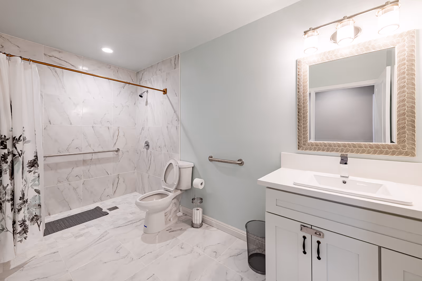 A clean, modern bathroom with white marble tile flooring and walls. The bathroom features a walk-in shower with a floral shower curtain, a white toilet with an open lid, a white vanity with a sink, a large decorative mirror above the sink, and a trash bin. There are grab bars installed near the toilet and in the shower area for accessibility.