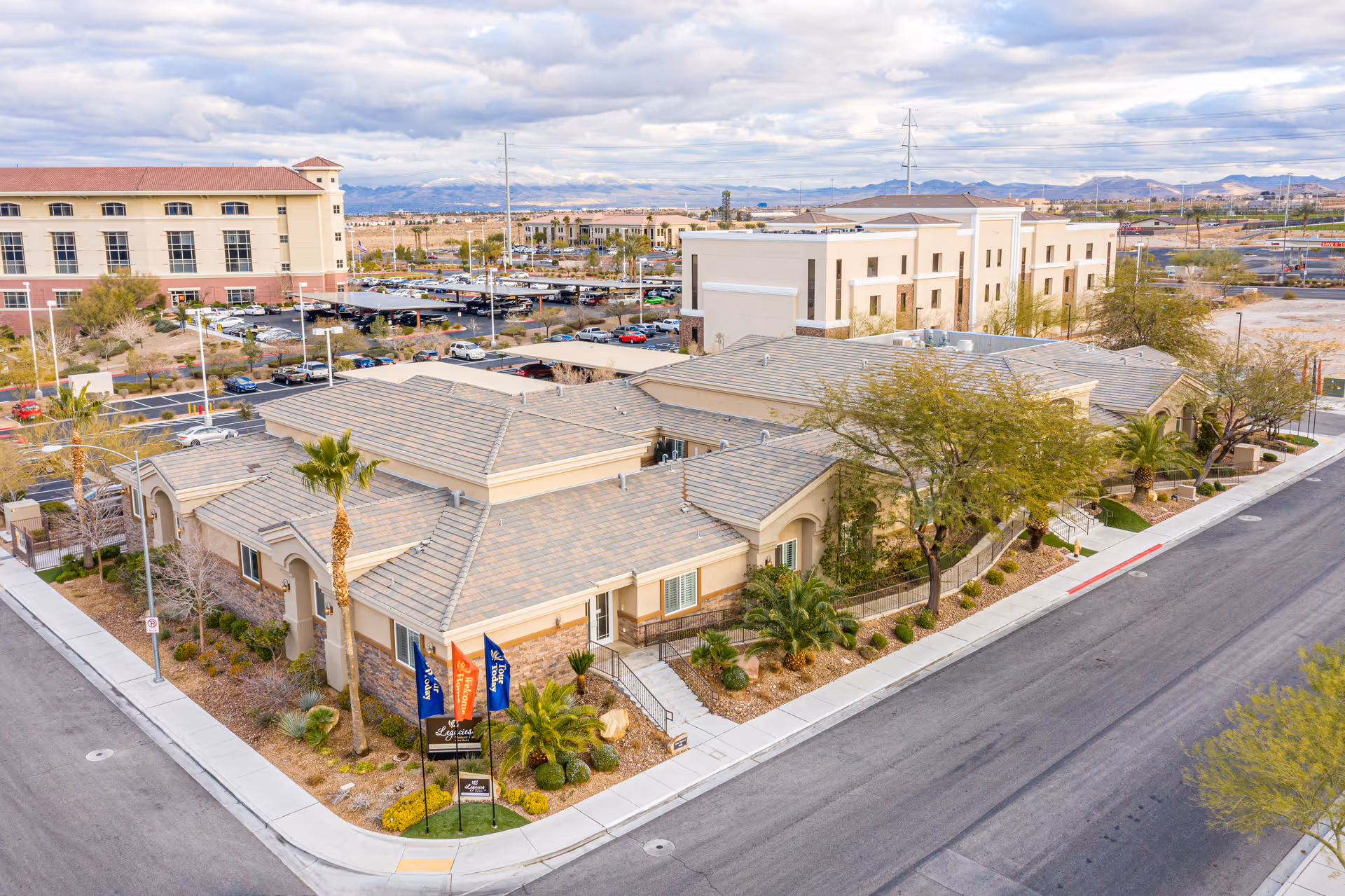 Aerial view of a single-story memory care facility building with beige walls and a tiled roof, surrounded by desert landscaping including palm trees and shrubs. The building is located at a street corner with sidewalks and a sign in front displaying the facility's name. In the background, there are parking lots, other buildings, and mountains under a cloudy sky.