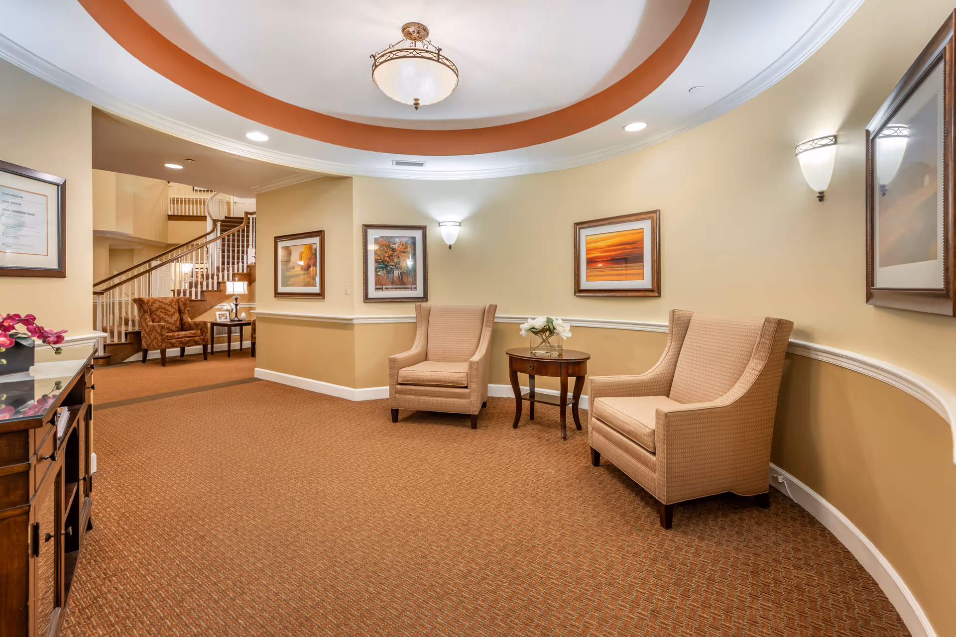 A cozy seating area in a senior living facility with two beige armchairs and a small round wooden table with a flower arrangement between them. The walls are painted in warm beige tones with white trim, decorated with framed landscape artwork. In the background, there is a staircase with wooden handrails and another armchair next to a small table with a lamp and picture frames. The ceiling features a circular recessed design with a central light fixture.
