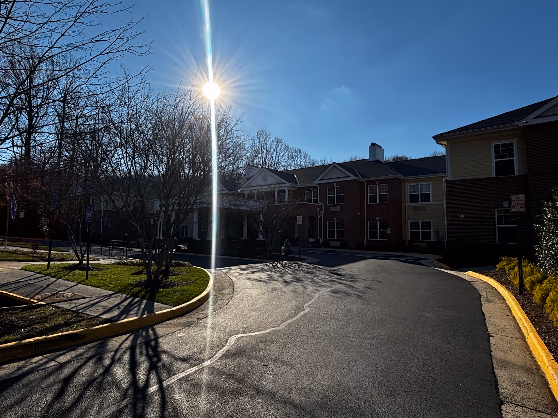 Exterior view of Chesterbrook Residences building on a sunny day with clear blue sky. The building has a brick and siding facade with multiple windows. Leafless trees and a curved driveway are visible in the foreground.