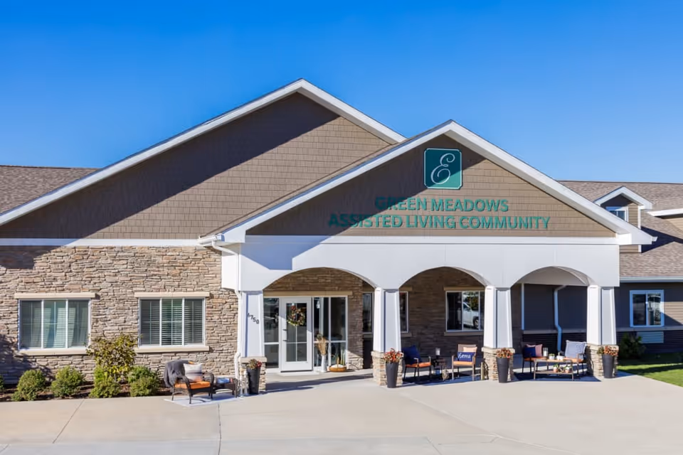 Front exterior view of Edencrest at Green Meadows assisted living community building with stone and siding facade, a covered entrance with white columns, outdoor seating, and clear blue sky.