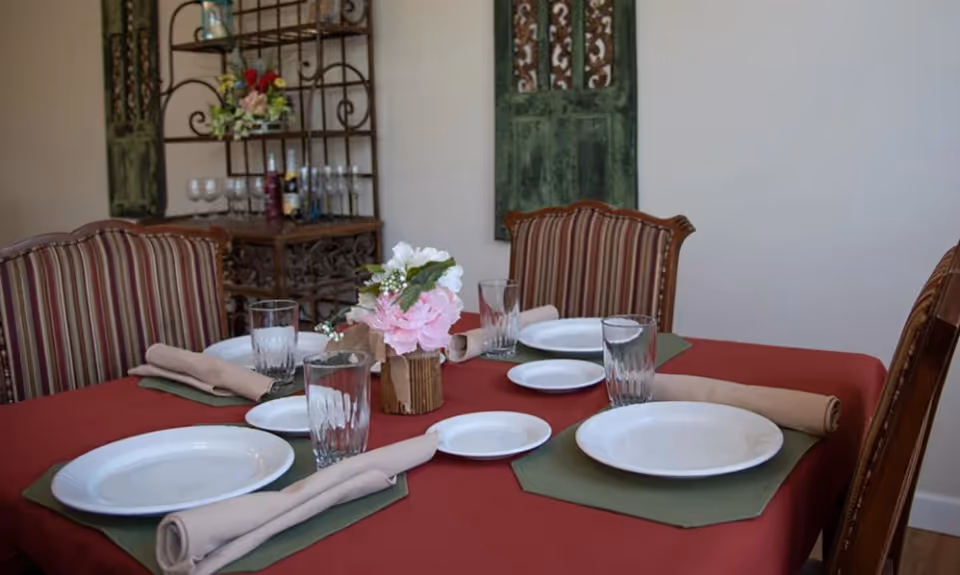 A dining table set for four with white plates, clear drinking glasses, beige rolled napkins on green placemats, and a small floral centerpiece in a wooden vase. The table has a red tablecloth and is surrounded by striped upholstered chairs. In the background, there is a decorative metal shelf with wine glasses and bottles, and two green decorative wall panels.