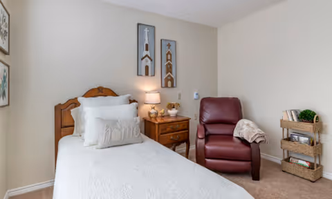 A cozy bedroom featuring a single bed with white bedding and decorative pillows, including one with the word 'home'. Next to the bed is a wooden nightstand with a lamp and small decorative items. A burgundy recliner chair with a beige throw blanket is positioned beside the nightstand. On the wall above the nightstand are two vertical framed artworks depicting church buildings. A small wicker shelf unit holds books and a plant. The room has beige walls and carpeted flooring.