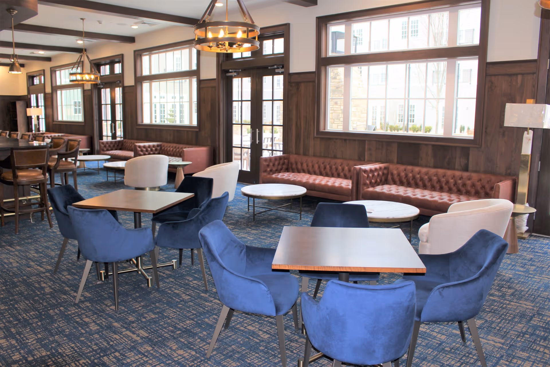 Bright communal lounge with blue upholstered chairs, tufted leather sofas, small tables and large windows.