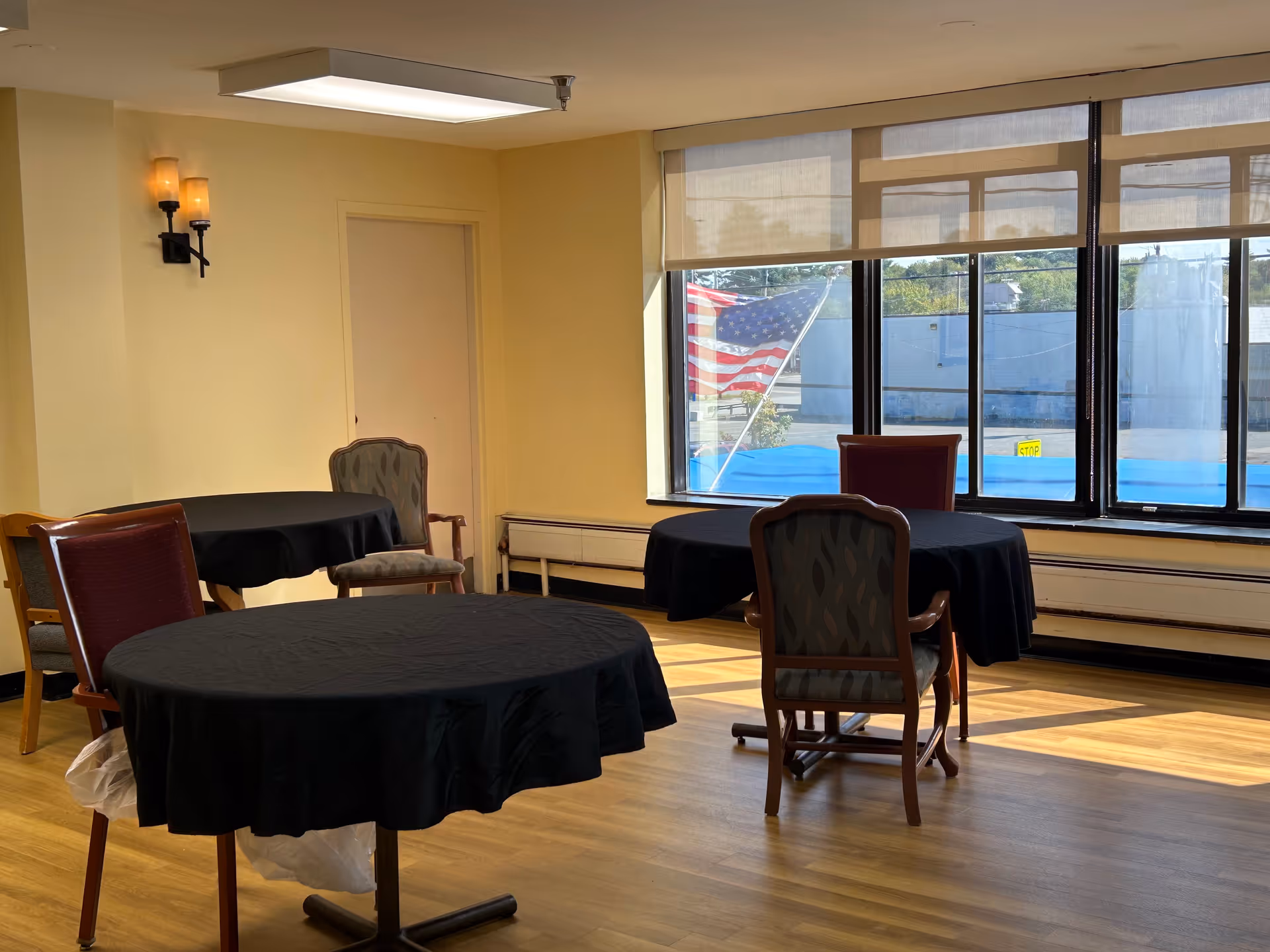 Sunlit common room with round tables covered in black tablecloths and chairs near large windows showing an American flag outside.