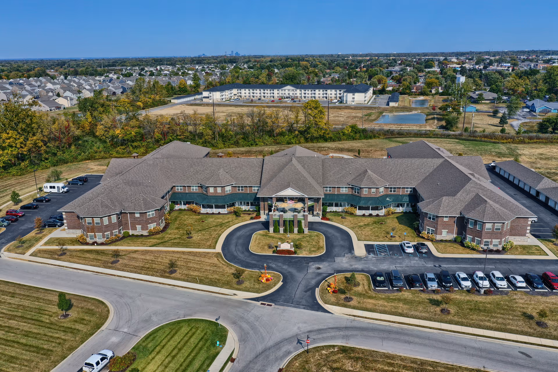 Aerial view of Magnolia Springs Southpointe, a large senior living facility with a U-shaped building surrounded by parking lots and landscaped lawns. The building has a central entrance with a covered driveway and multiple wings extending on either side. The surrounding area includes residential neighborhoods, trees, and open fields under a clear blue sky.
