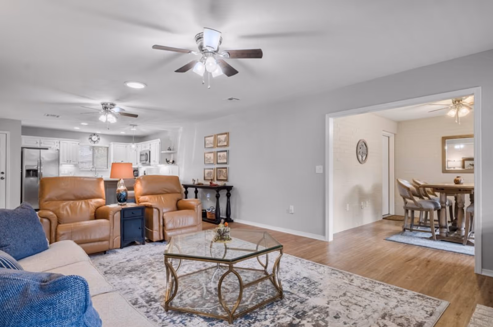 A bright and spacious living room with two brown leather recliners, a beige sofa with blue cushions, a glass coffee table with a decorative item, and a patterned area rug. The room has ceiling fans with lights and an open view into a kitchen with white cabinets and stainless steel appliances. Adjacent to the living room is a dining area with a wooden table and chairs, a wall clock, and a mirror.
