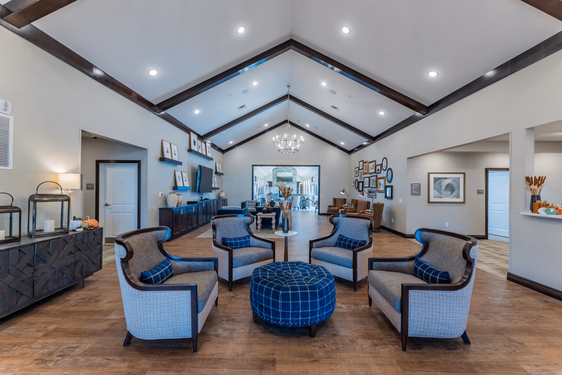 A spacious senior living facility common area with a high vaulted ceiling featuring dark wooden beams and recessed lighting. The room has a seating arrangement of four cushioned armchairs with blue plaid pillows around a round blue plaid ottoman. The walls are decorated with framed pictures and shelves holding decorative items. There is a dark wooden sideboard with lanterns and small pumpkins on the left side. The flooring is wood, and the room has a warm, inviting atmosphere.