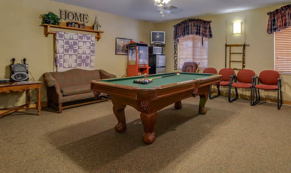 A recreational room with a pool table in the center, a small couch against the wall with a decorative quilt and a shelf above it, a popcorn machine and a small TV on a stand in the corner, several red chairs lined up against the wall, and windows with plaid valances. The room has beige walls and carpeted floor.
