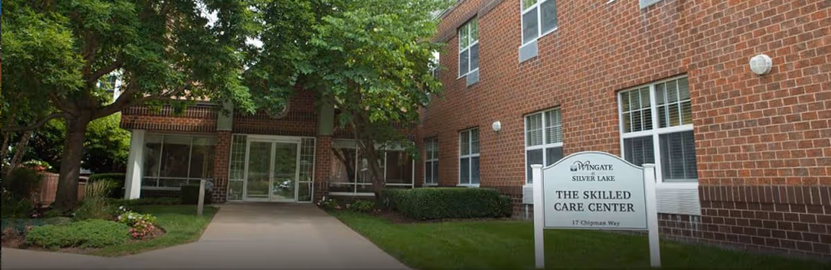 Exterior view of Wingate at Silver Lake Skilled Care Center showing a brick building with multiple windows, a glass entrance door, surrounding greenery including trees and bushes, and a sign with the facility name and address.