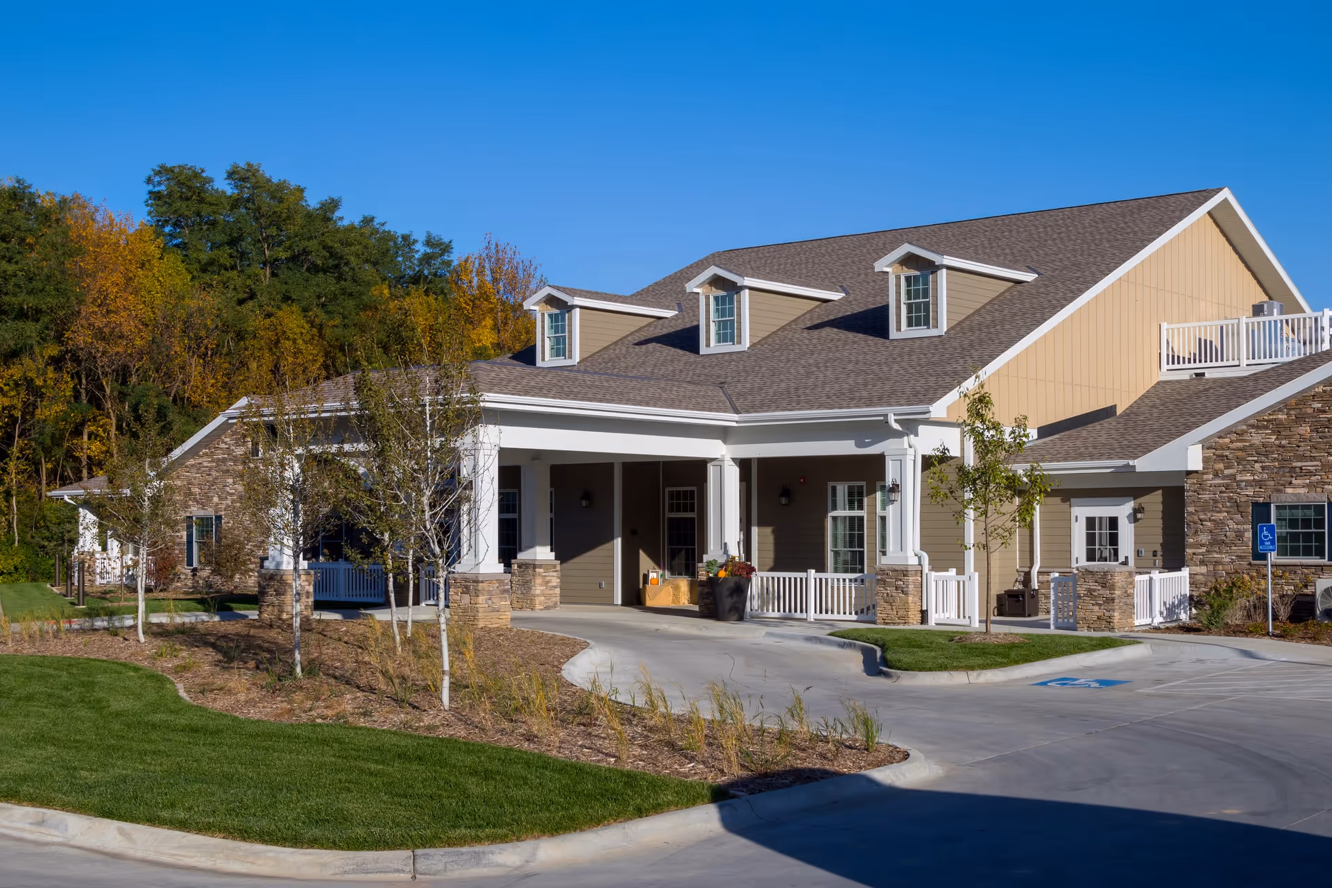 Exterior view of a single-story senior living facility building with a covered entrance, beige siding, stone accents, and a well-maintained lawn and landscaping under a clear blue sky.