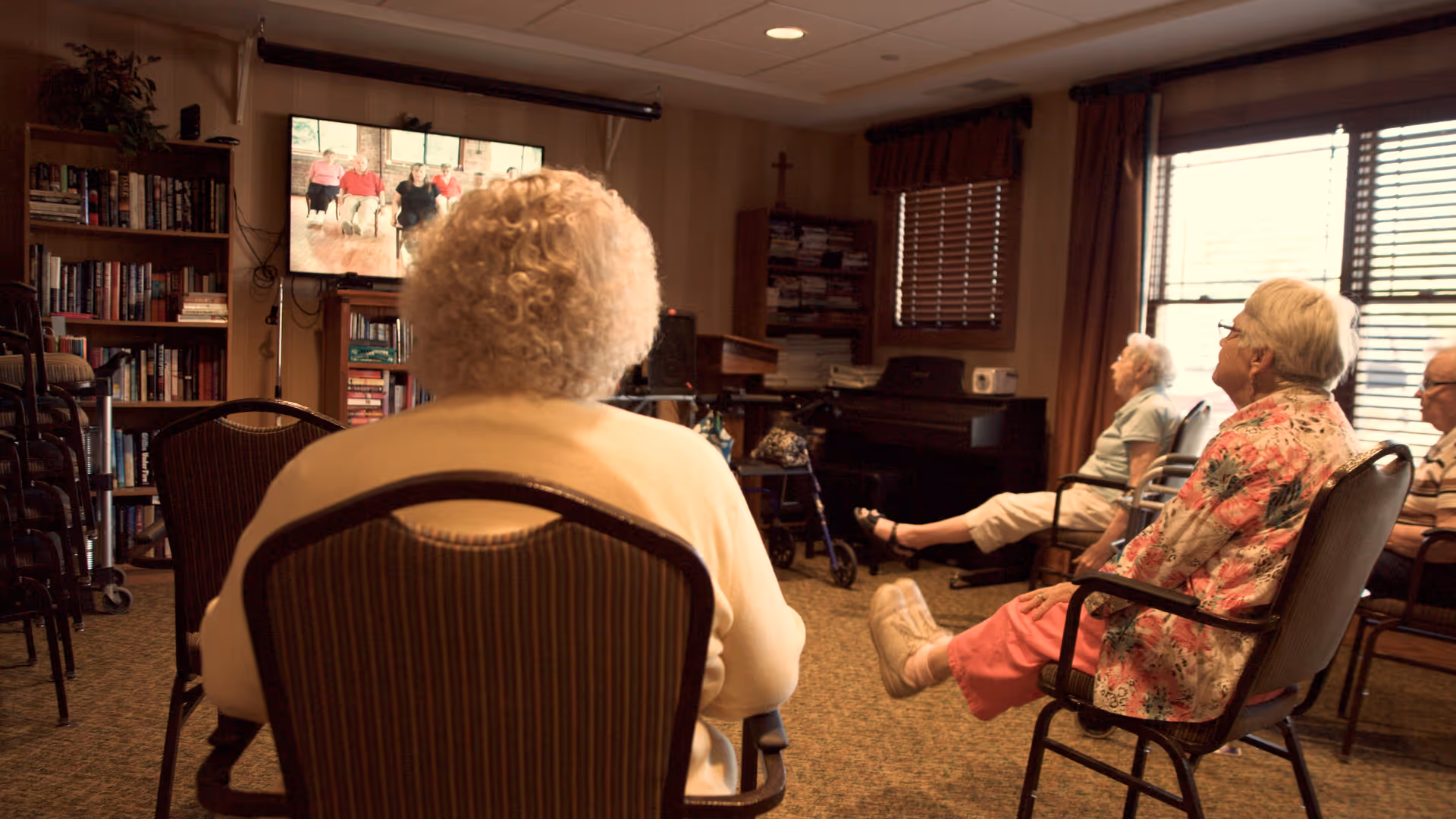 A group of elderly people seated in chairs in a common room watching a television screen. The room has bookshelves, a piano, and large windows with blinds allowing natural light to enter.