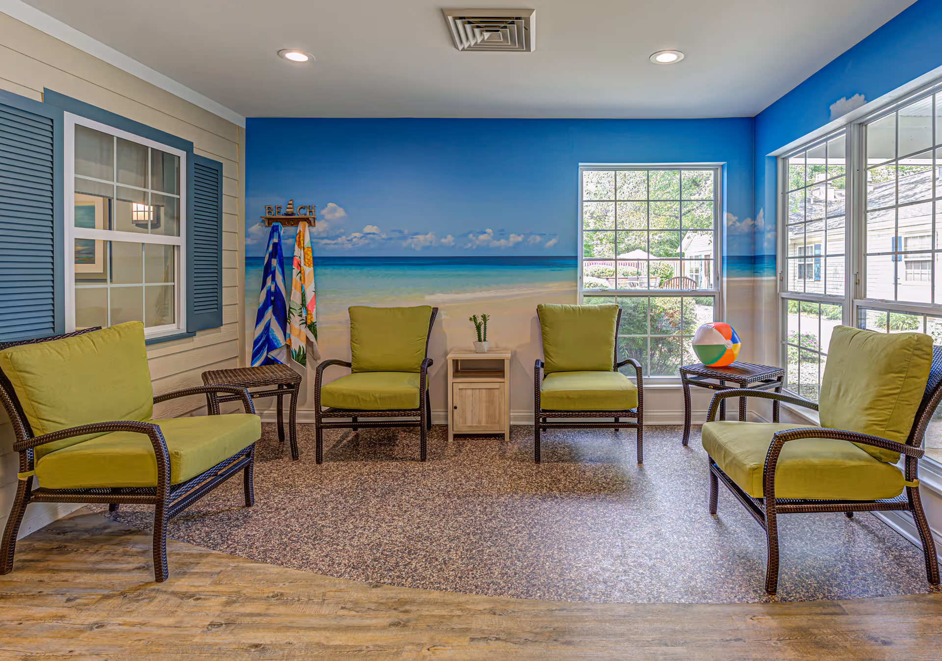 A bright room with four green cushioned chairs arranged around two small tables. The walls feature a beach-themed mural with a blue sky, ocean, and sand. There are two large windows letting in natural light, a small wooden side table with a plant, a colorful beach ball on one table, and a coat rack with beach towels hanging on it.