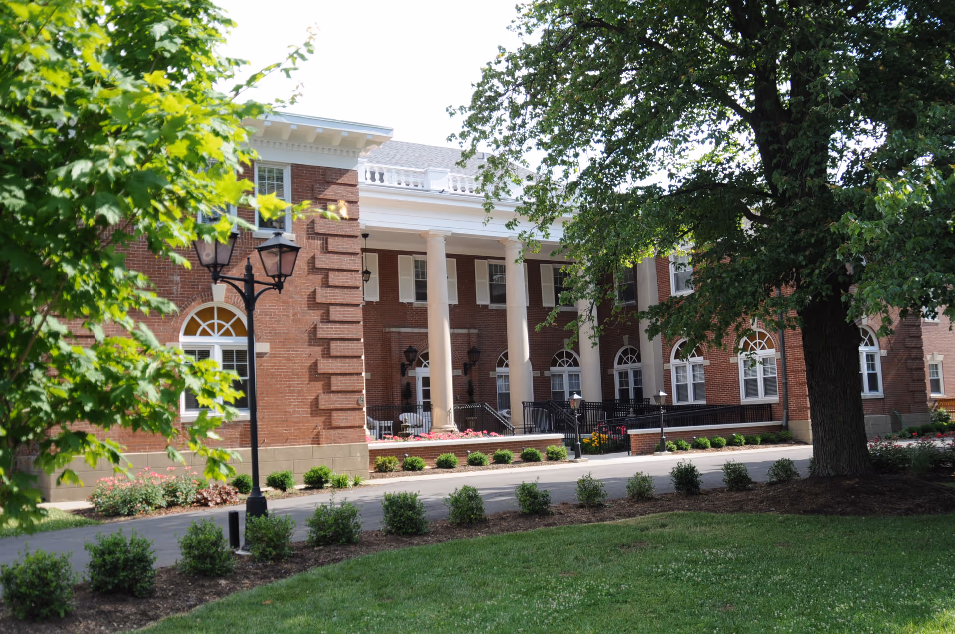 Exterior view of a senior living facility building with red brick walls, white columns, and multiple windows. The foreground features a well-maintained lawn, small bushes, a large tree, and a black street lamp.