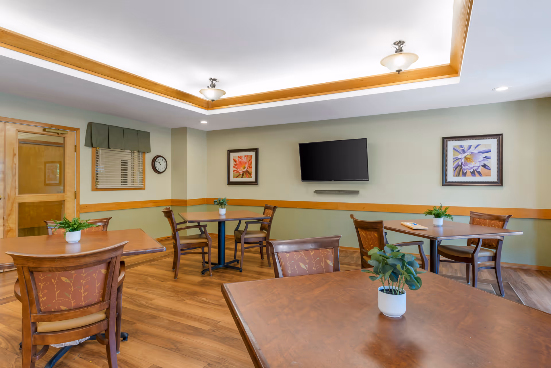 Well-lit common dining room with several wooden tables and chairs, potted plants, a wall-mounted TV, and framed artwork.